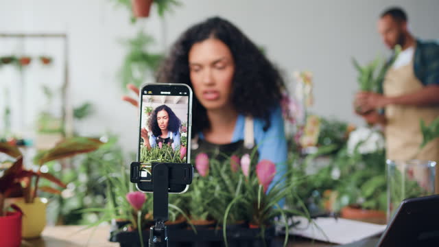 Smiling female social media blogger designer florist shooting new video on smartphone, talking about gardening and planting. Female gardener streaming live video, conference calling or making creative web training lesson in flower shop.
