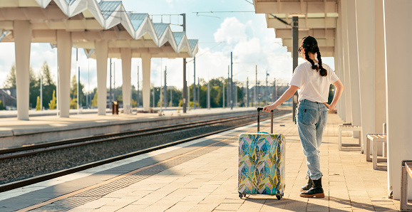 Back view of woman holding her luggage and waiting departure. Train station is in background. Copy space. Web banner of modern travel and vacation.