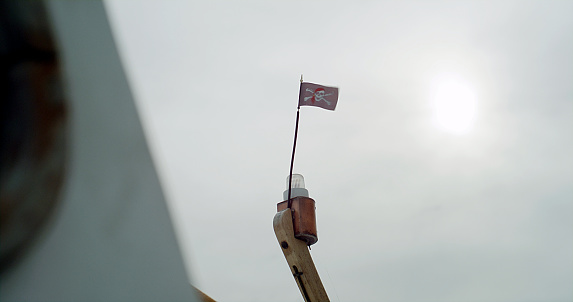 Pirate flag waving on the wind on top of ship's mast. Clear sky and bright sun