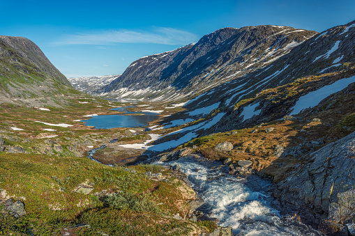 The snow capped Breiddalen Valley at Jotunheimen National Park in Norway, along the route to Geiranger, during a summer day. A cabin rests near the base of a mountain.