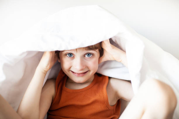istock Little boy sitting and smiling under white blanket in bedroom. 1755711062