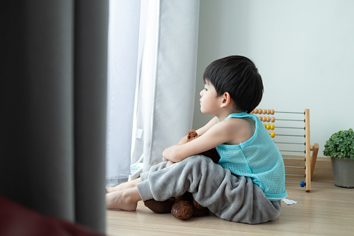 Sad Asian boy sits with his head down and looks out the door at his favorite doll.