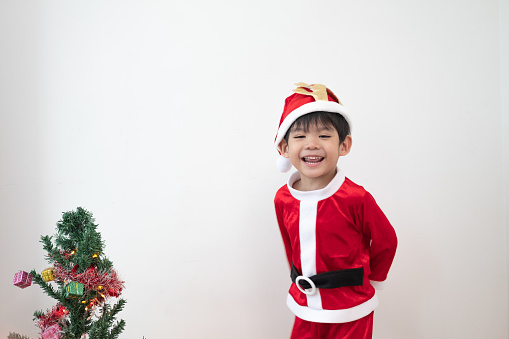Asian boy wearing Santa costume Standing and playing near A fun Christmas tree on white background.