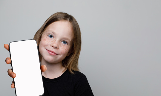 Cute little child showing smartphone with blank empty white screen display on gray background