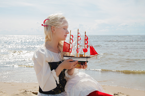 Beautiful young adult woman holding small yacht with red sails on sea beach