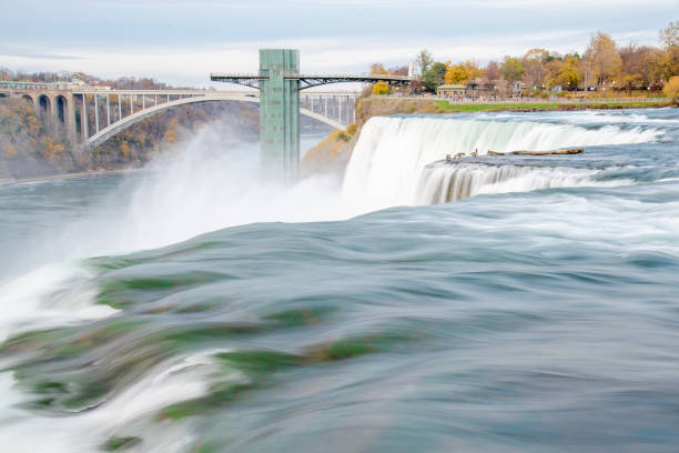 Long exposure American Falls waterfall cascade with gooses near Niagara Falls Observation Tower and Rainbow International Bridge, view from Luna Island, autumn leaves foliage sunset. Travel tourist