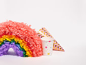Festive birthday decoration isolated on white background. A colorful rainbow pinata, a nice paper cup and a holiday hat. Copy space for text.