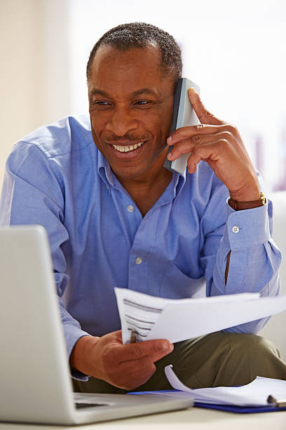 Mature African man working from home Image of  mature man holding few papers looking at laptop and making a phone call cordless phone stock pictures, royalty-free photos & images