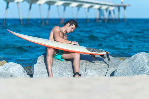 A Serious young man cleaning surfboard on sunny beach