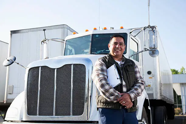 Hispanic truck driver (40s) standing in front of semi-truck with clipboard.