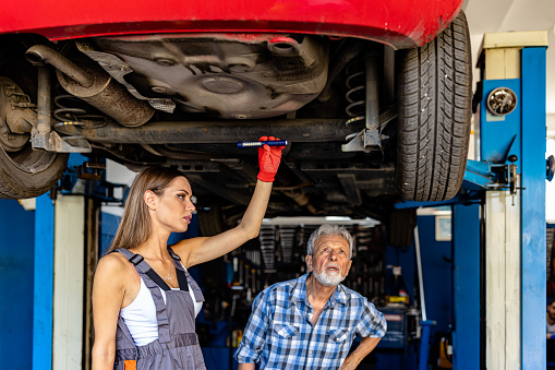 An Experienced Car Mechanic is Standing Under the Car in a Car Service and Fixing the Problems with the Assistance of his Young Female Colleague.