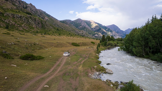 A white SUV overcomes a mountain route along a gorge along the river. Green fields with cliffs. Steep cliffs and big rocks on the road. A sharp ascent and descent with a view of hills. Christmas trees