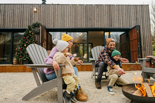 Photo of a cheerful family of four, roasting marshmallows by the campfire on a chilly autumn day, as they are spending extended weekend in a log cabin