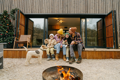 Photo of a cheerful family of four, roasting marshmallows by the campfire on a chilly autumn day, as they are spending extended weekend in a log cabin