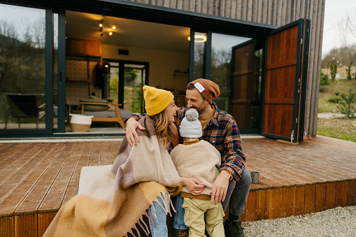 Photo of a smiling, cheerful family who enjoy each others company during a prolonged weekend in front of a cabin house - their rental home for the weekend