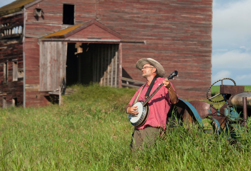 A man playing a banjo in a rustic rural scene. Abandoned grain elevator in distance. Horizontal colour image. White male. Early 40s. Prairie setting.