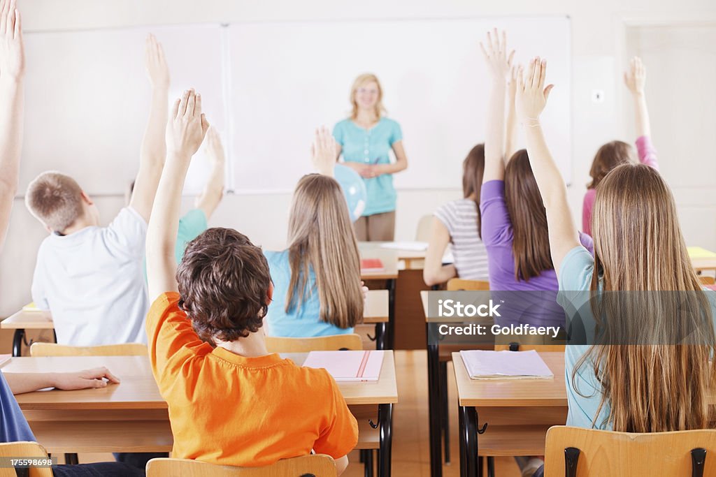 Pupils in classroom responding to the teacher's question. "Rear view of students in bright classroom responding to the teacher's question, raising their arms up." Instructor Stock Photo