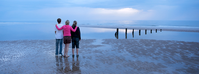 Peaceful scene of a mum and her two teenage children on a beach, watching as the last light of the sun goes down at twilight.