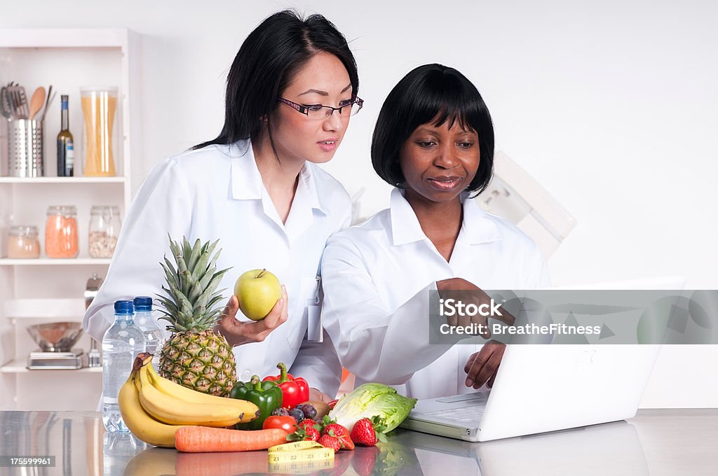 Dietitians Dietitians discussing some information on a laptop with a pile of fruit and vegetables next to them.See more dietitians here: Nutritionist Stock Photo