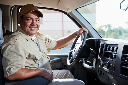 A Hispanic worker in his 40s wears a beige uniform and drives a commercial van.  The man is smiling and facing the camera, with his left hand on the steering wheel.  He is wearing a brown baseball cap, and trees and a building are in the background.