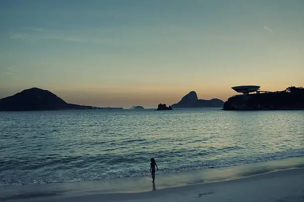 "A girl watching the sunset from the beach. On the right, a building lika an UFO.NiterAi, Rio de Janeiro - Brazil.Camera: Canon EOS 30D.Post-production: digital cross-processing."