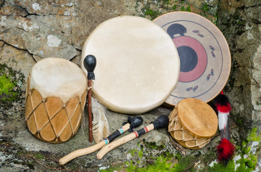 Four, Native American Frame Drums, with Drum Sticks, Brightly colored feathers on a natural stone wall background.