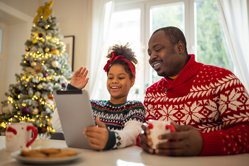 Father and daughter drinking tea and eating cookies. They are wearing Christmas sweaters and having video call with family.