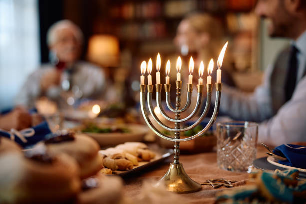 Lit menorah on dining table with Jewish family celebrating Hanukkah in the background. Lit candles in menorah during Hanukkah celebration. The family is in the background. hanukkah stock pictures, royalty-free photos & images