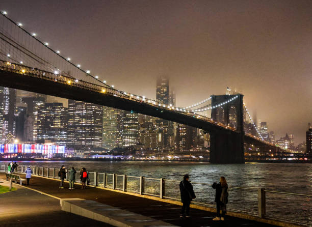 Multiple groups of people strolling on the Brooklyn Bridge Park boardwalk on a foggy night. The walkers were looking at the bridge and Manhattan skyline, which was covered in a mystical fog.