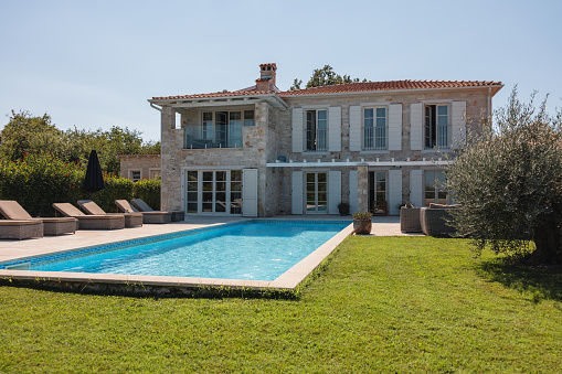 A beautiful Istrian villa with a large pool in the backyard. The sun is shining and illuminating the pool and the grass in the backyard.