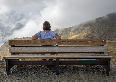 A curvy young Caucasian girl enjoying a well-deserved rest sitting on a wooden bench relaxed thoughtfully and looking at the landscape from the top of Tristaina mountain in Andorra