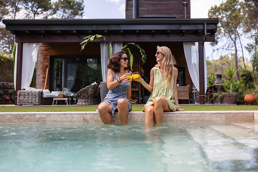 Attractive Caucasian female friends enjoying their time by the pool on a hot summer day. They are sitting by the edge of the pool and toasting with their glasses of orange juice. They look happy and are smiling. The women are wearing cute summer dresses.