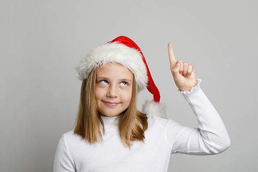 Christmas portrait of funny child girl in Santa hat pointing up on white background
