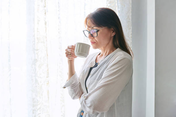 istock Serious middle-aged woman looking out window, with cup in hands 1756127012