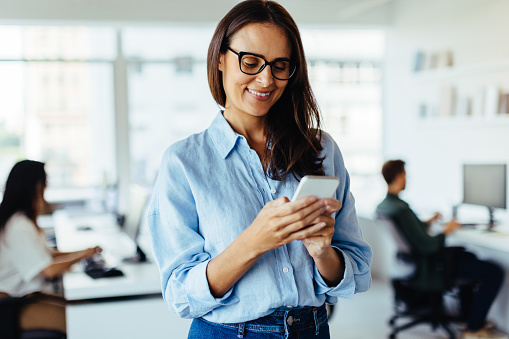 Business woman using a smartphone while standing in an office. Creative woman updating her professional social media page.