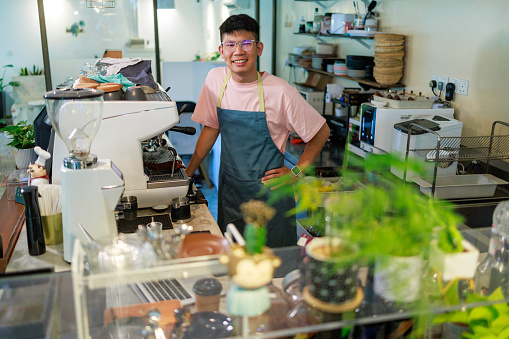 Young Asian barista standing confidently in front of the counter. The male barista is the business owner of a small cafe, appearing cheerful and passionate about his work. This scene exemplifies the concept of a small business