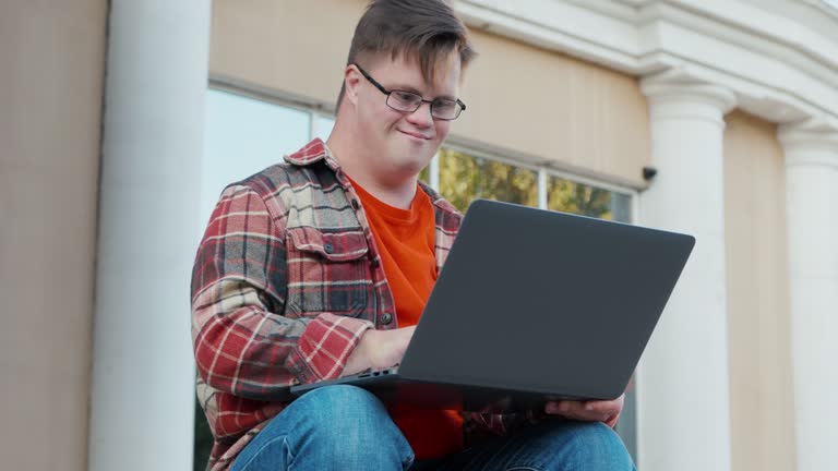 Young man with down syndrome in checkered shirt using laptop working online outdoors in the city. Guy with disability working remote online as freelancer