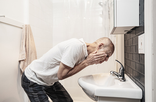 Senior man washing his face after shaving in the morning in his bathroom. He is dressed in white t-shirt and pyjama bottoms. Interior of private home in Toronto, Canada.