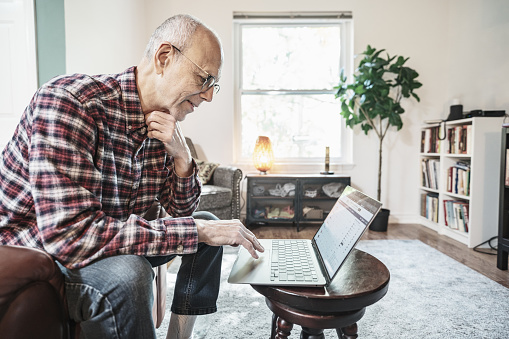 Senior man using laptop in his living room. He is dressed in casual clothes. Interior of private home in Toronto, Canada.