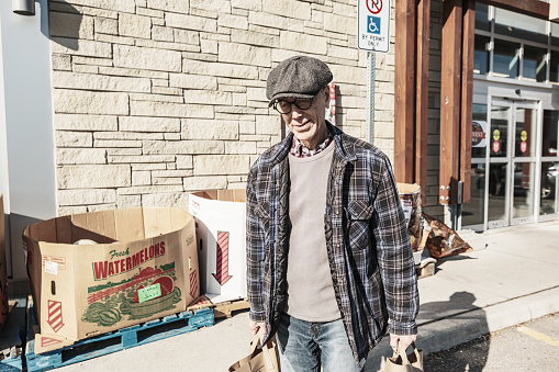 Senior man leaving grocery store after shopping. He is dressed in casual clothes. Exterior of parking lot on the street  in Toronto, Canada.