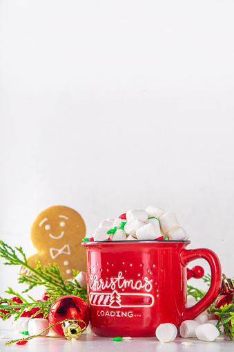 Funny cute gingerbread man with hot chocolate, cappuccino or latte. Festive Christmas cocoa mug with marshmallow and cookie, with Christmas tree, decor, on white bokeh background copy space