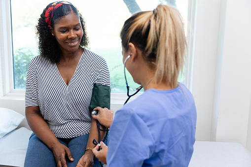 A mature adult female patient smiles as an unrecognizable nurse checks her blood pressure during a visit to the doctor.