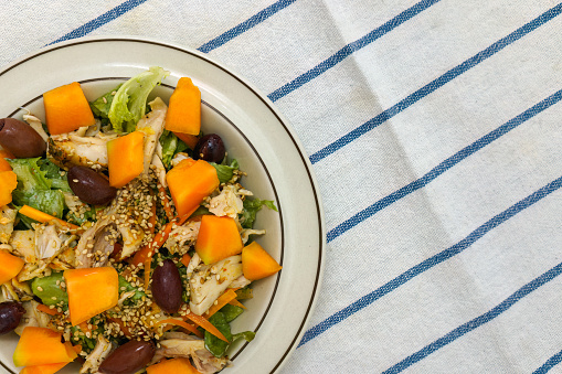 Fresh chicken salad with kalamata olives, cantaloupe melon and sesame seeds. Top view on white kitchen towel with blue stripes.