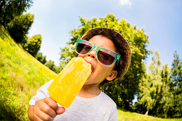 Little boy licking orange popsicle in park Little latino child enjoying a popsicle in park candy in mouth stock pictures, royalty-free photos & images