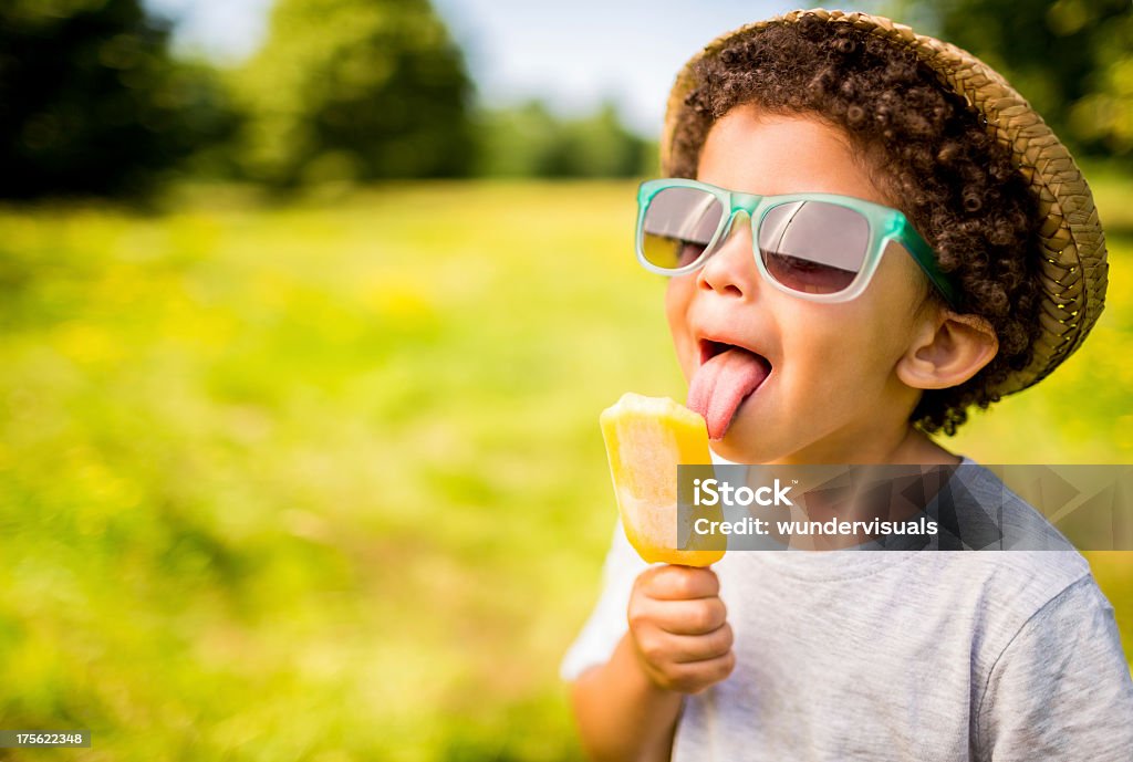 Boy in sunglasses and hat eating popsicle outdoors The tongue of a young boy with curly hair is touching an orange frozen treat.  He is wearing a gray t-shirt, a straw hat and blue rimmed glasses.  The background is blurry and consists of an open field of green grass with trees and bushes in the distance. Child Stock Photo