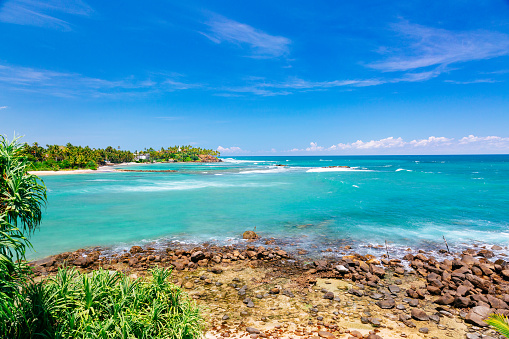 A beautiful tropical beach with a rocky island covered in greenery in the background. The sky is a bright blue with white clouds. The water is a clear turquoise color and the sand is white. The photo is taken from the shore looking out towards the ocean.