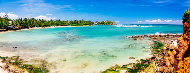 A panoramic photo of a beautiful tropical beach with clear blue water and white sand. The beach is lined with palm trees and there are a few small buildings visible. The sand is white and there are some rocks scattered along the shore. The ocean is a beautiful turquoise color and the water is clear. The photo was taken from a high vantage point, looking down on the beach.