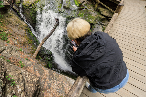 Mature blonde woman taking photos of Bushkill Falls in Pennsylvania from wooden bridge trail at Pocono Mountains in Pennsylvania. Top view