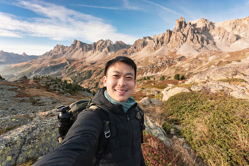 Happy young asian backpacker man taking selfie on the top of mountain in Valley Claree with iconic mountain peak in autumn at French Alps, Hautes Alpes, France