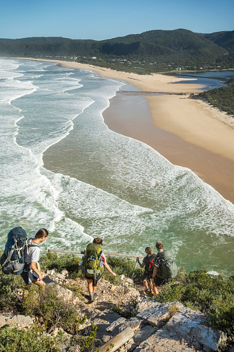 Rear view of group of hikers walking down the mountain trail towards the beach on a summer day